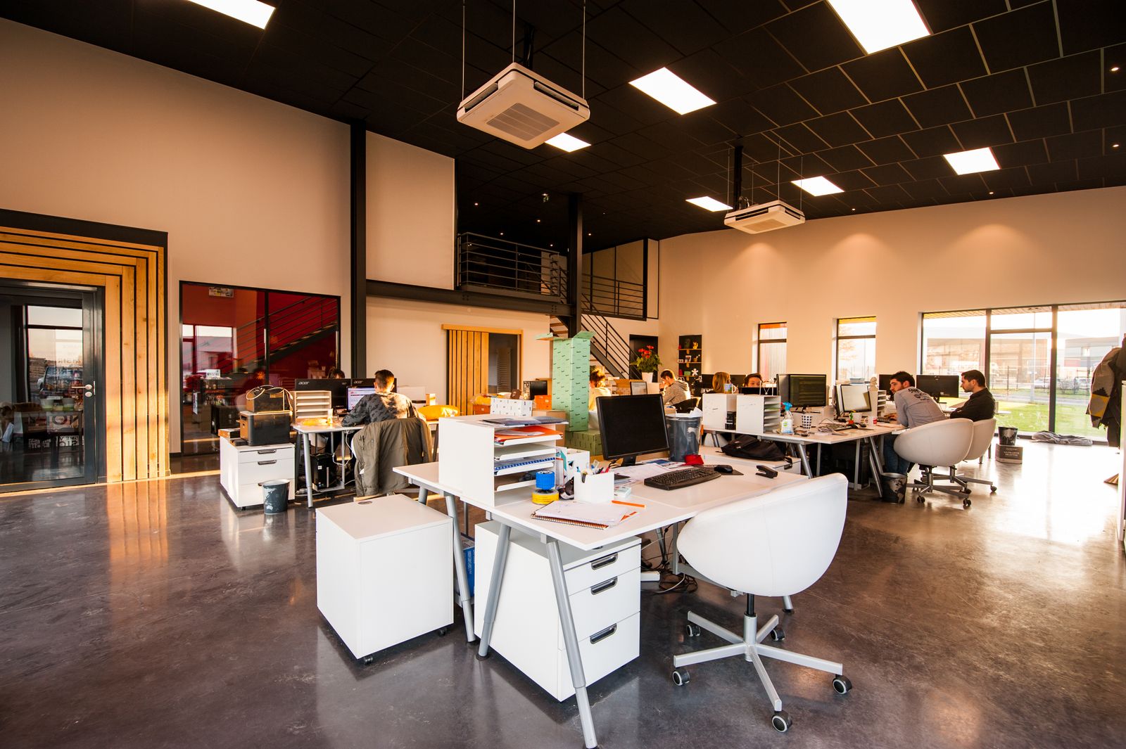 An open-plan tech office mid-afternoon: people working at desks with monitors, a mezzanine above and windows along one wall.